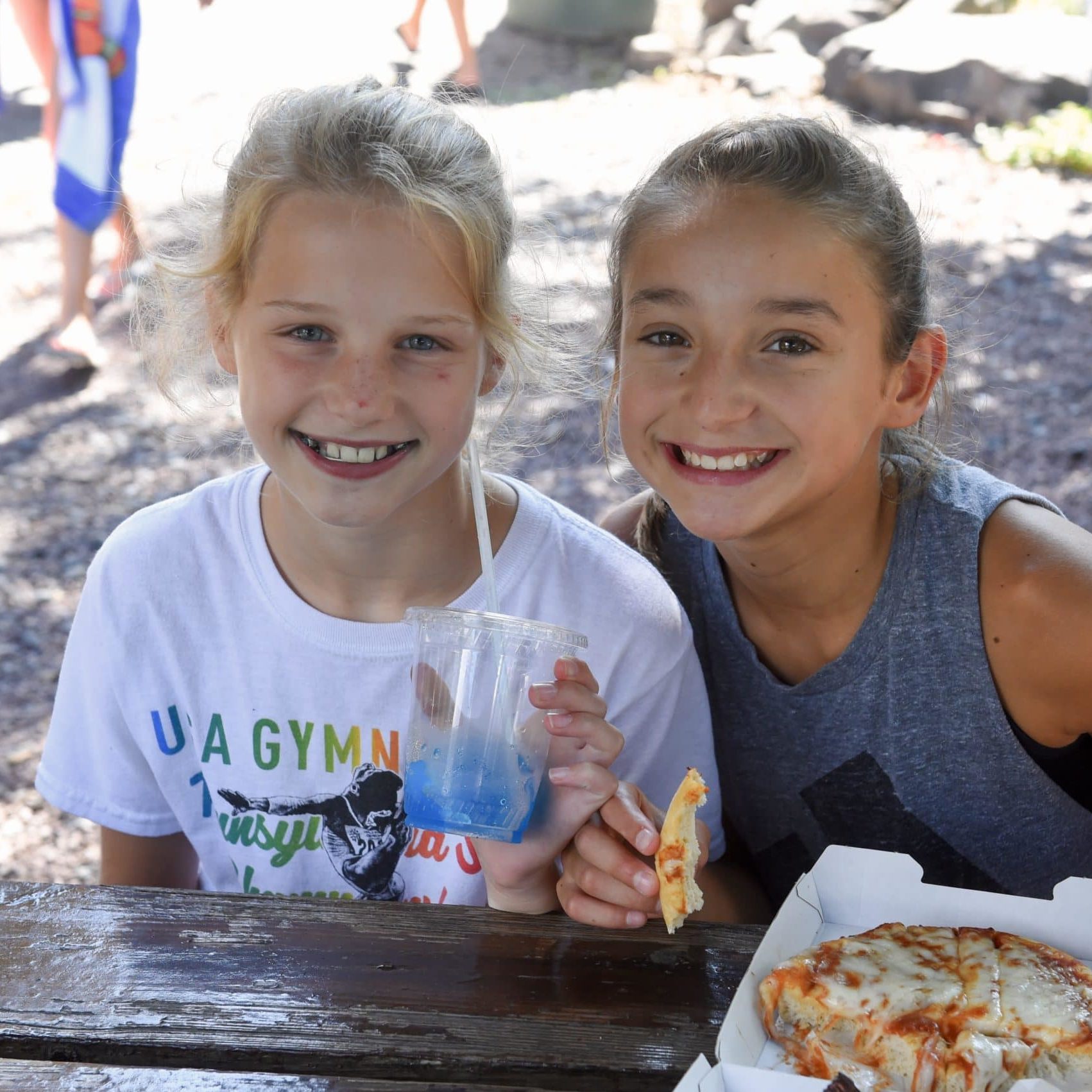 Two campers pose for a photo during lunch. One girl has a blue beverage and one camper has a pizza in front of her.