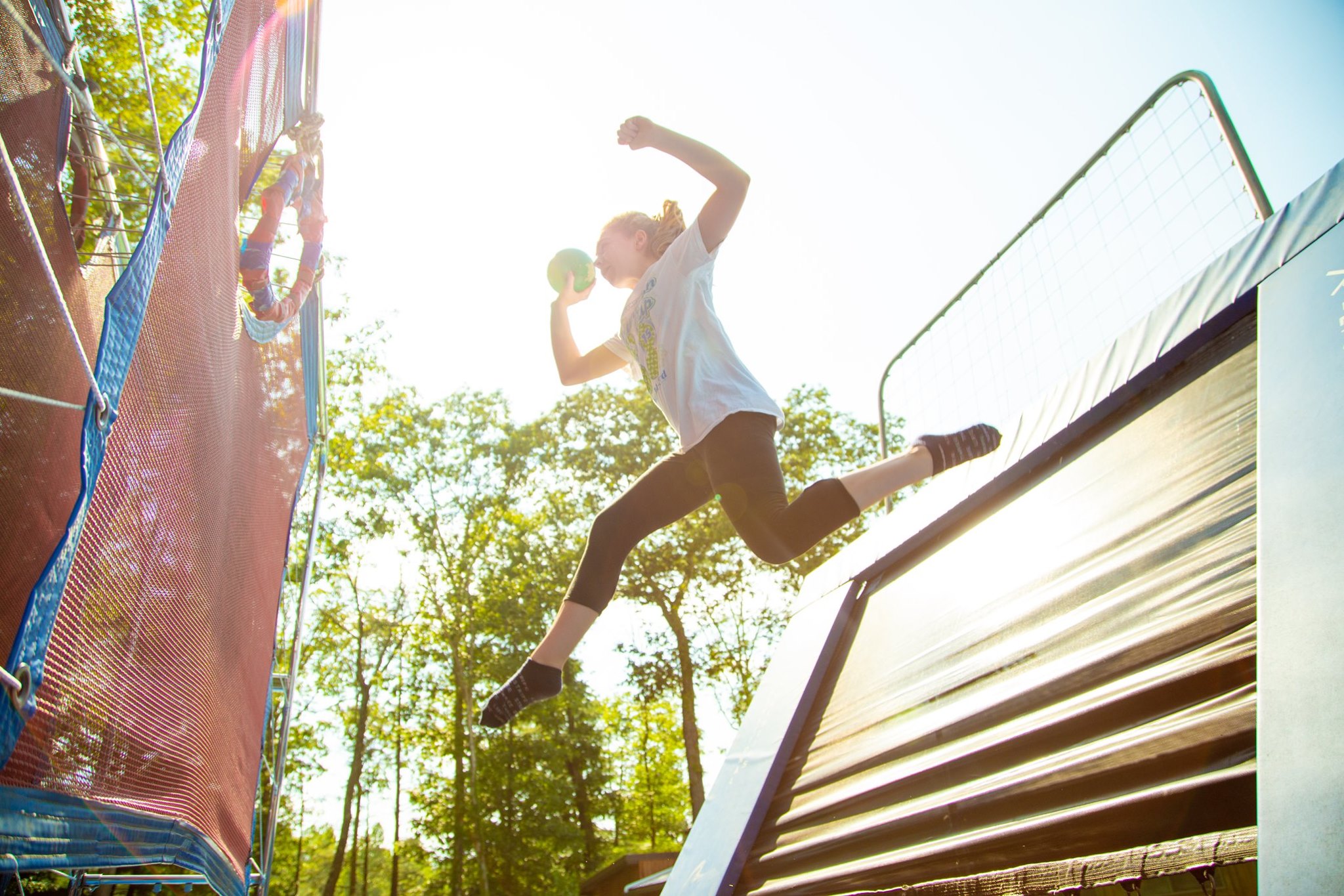 A girl jumps in the air with a ball in her hand. There are trees and the sun behind her.