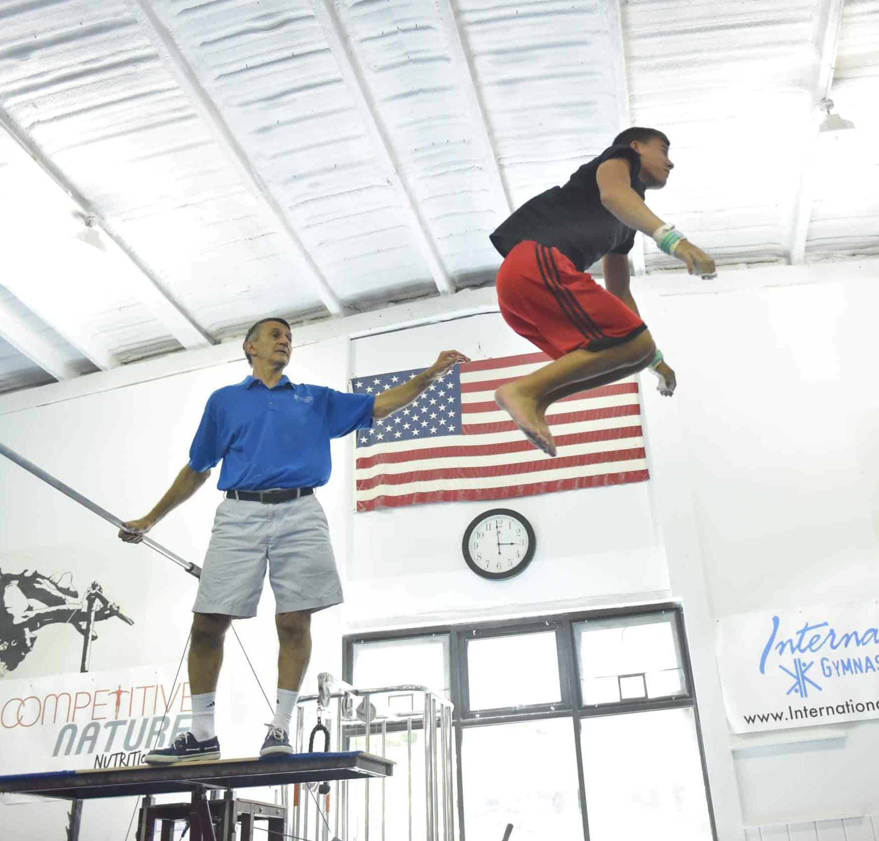 A camper does a gymnastics trick in the air as a staff member watches.